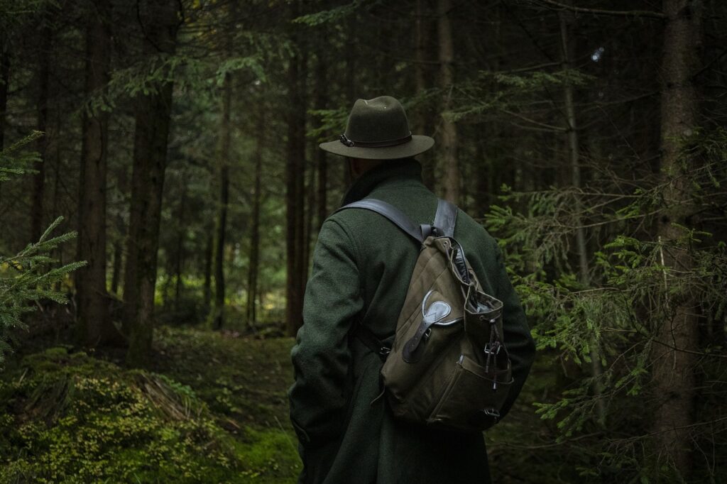 Wanderer in einem saftig grünen, dunklen Wald, umgeben von hohen Bäumen und dichtem Unterholz. Das dichte Blätterdach lässt nur vereinzelte Lichtstrahlen durch, die eine mystische Atmosphäre schaffen.
