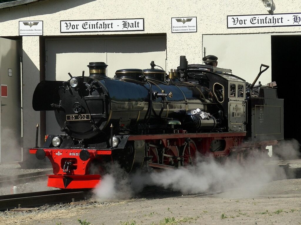 Eine Dampflok, die aus einem Bahnhof fährt und dabei kräftigen Dampf ausstößt. Der Zug ist von Rauch und Dampf umhüllt, während die Eisenbahnstrecke in den Horizont führt und eine nostalgische Atmosphäre erzeugt.